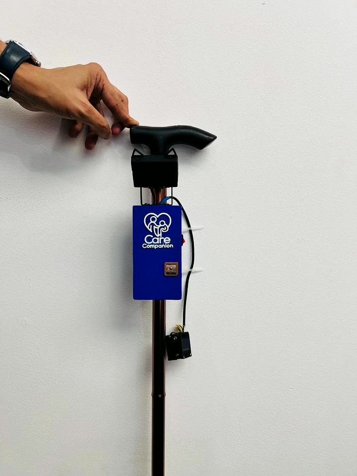 A hand resting on the black T-handle of a rose-gold folding walking cane. The blue CareCompanion enclosure is fixed to the shaft just below the handle, the HuskyLens camera on its bracket above it, and the display module hanging on a lead below.