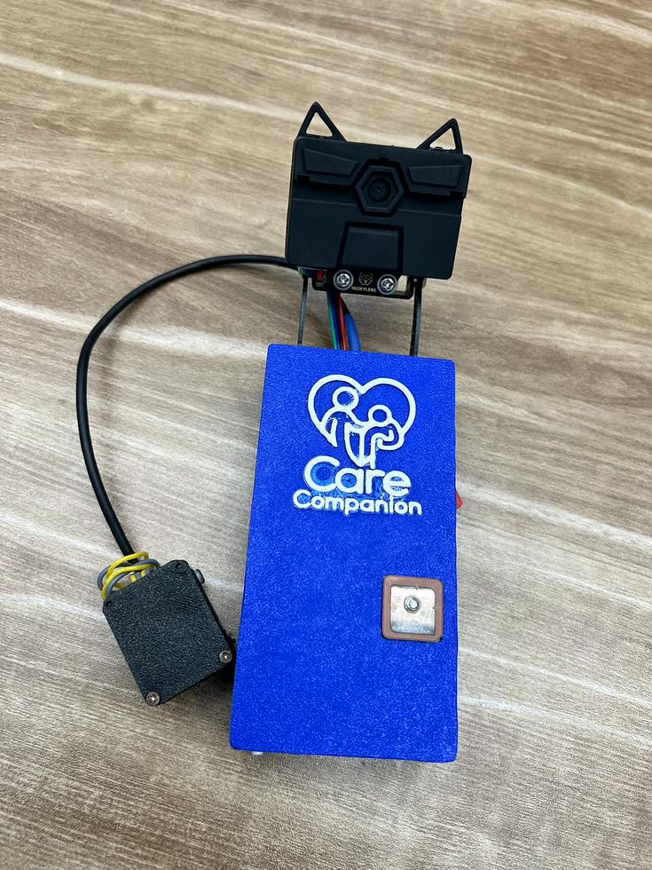 The device on a desk, off the cane: the blue enclosure carrying the team's logo and the GPS patch antenna, the HuskyLens camera above it on its bracket, and the display module to one side.