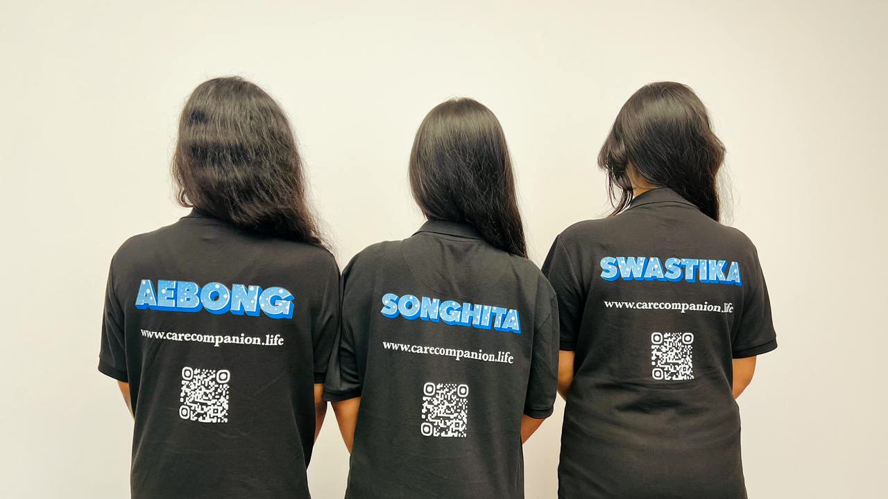The three of them from behind, showing the backs of the team shirts: AEBONG, SONGHITA and SWASTIKA, each above the address www.carecompanion.life and a QR code.